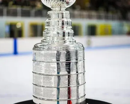 An image of the hockey Stanley Cup; a silver multi-teered metal trophy in the shape of a cup. It sits on a black table with a ice rink background.