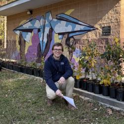 A person kneeling in front of a row of potted plants outside of a building with a colorful mural.