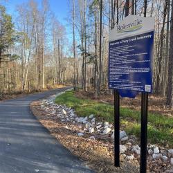 Asphalt walkway with brown leaves and trees and a blue rules sign