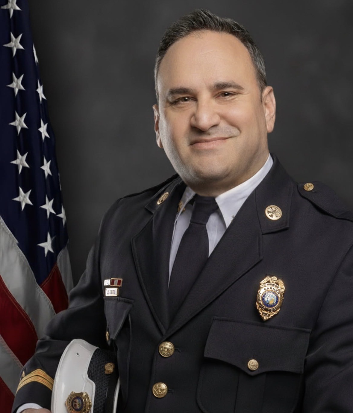 Man standing in front of black background with an american flag to the left and he's wearing a black suite